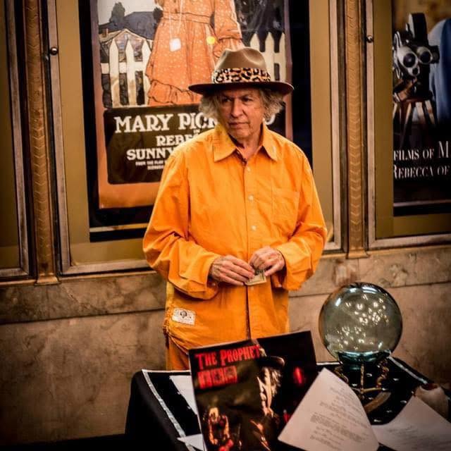 Stephen Kalinich in an orange shirt and hat, standing beside a display of books and film memorabilia